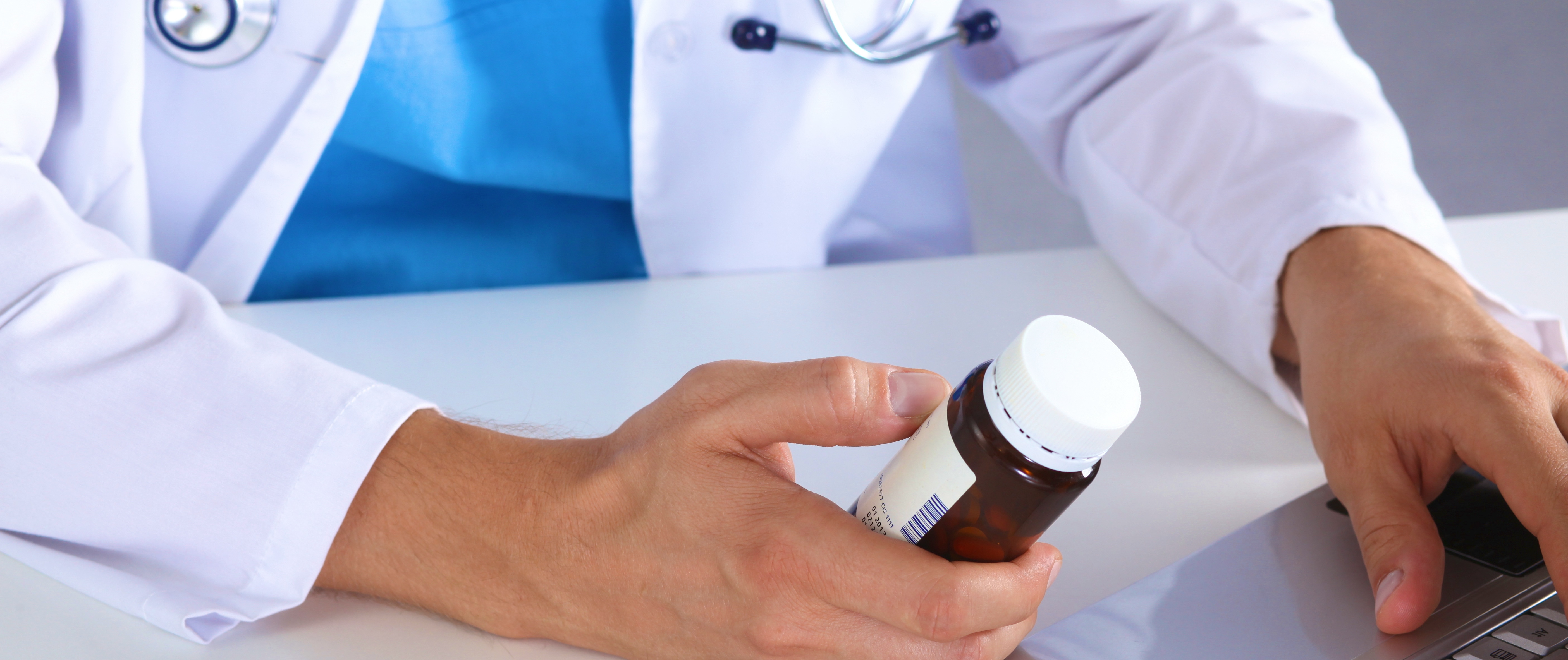 A person in a medical jacket holding a box of pills typing on a laptop